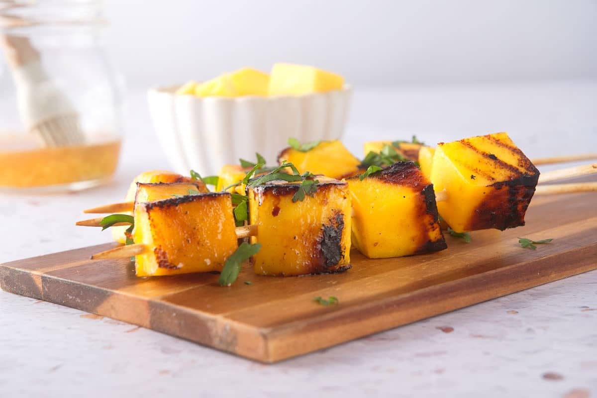 Grilled mango skewers on wooden serving board with fresh cilantro.