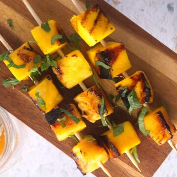 Grilled mango skewers on wooden serving board with fresh cilantro.