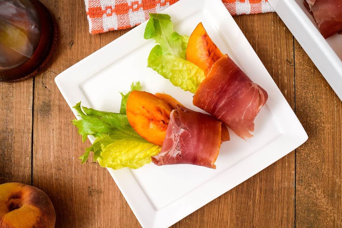 Grilled Peaches Wrapped in Proscuitto on a porcelain appetizer plate, balsamic dressing and peaches in the background.