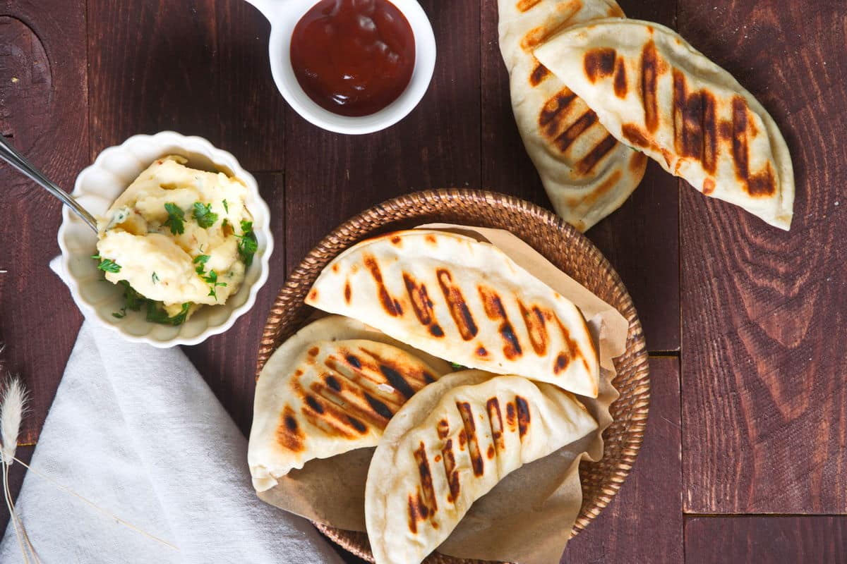 Top view of grilled pierogies in wooden basket. BBQ sauce and filling on the side.