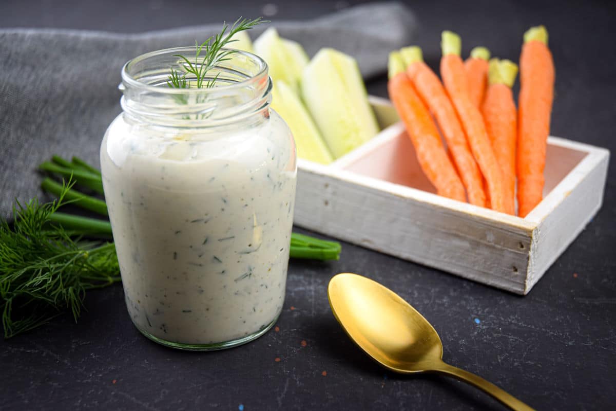 Homemade ranch dressing in a jar with fresh dill, carrots, cucumbers and a golden spoon on the side.