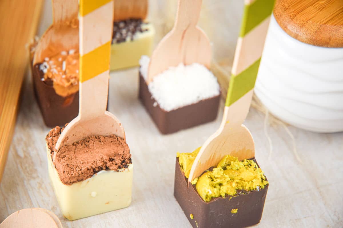 Hot chocolate spoons with toppings on light brown background.