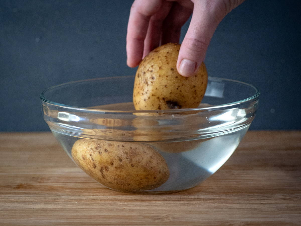 Potatoes in a bowl of water, on wooden cutting board.