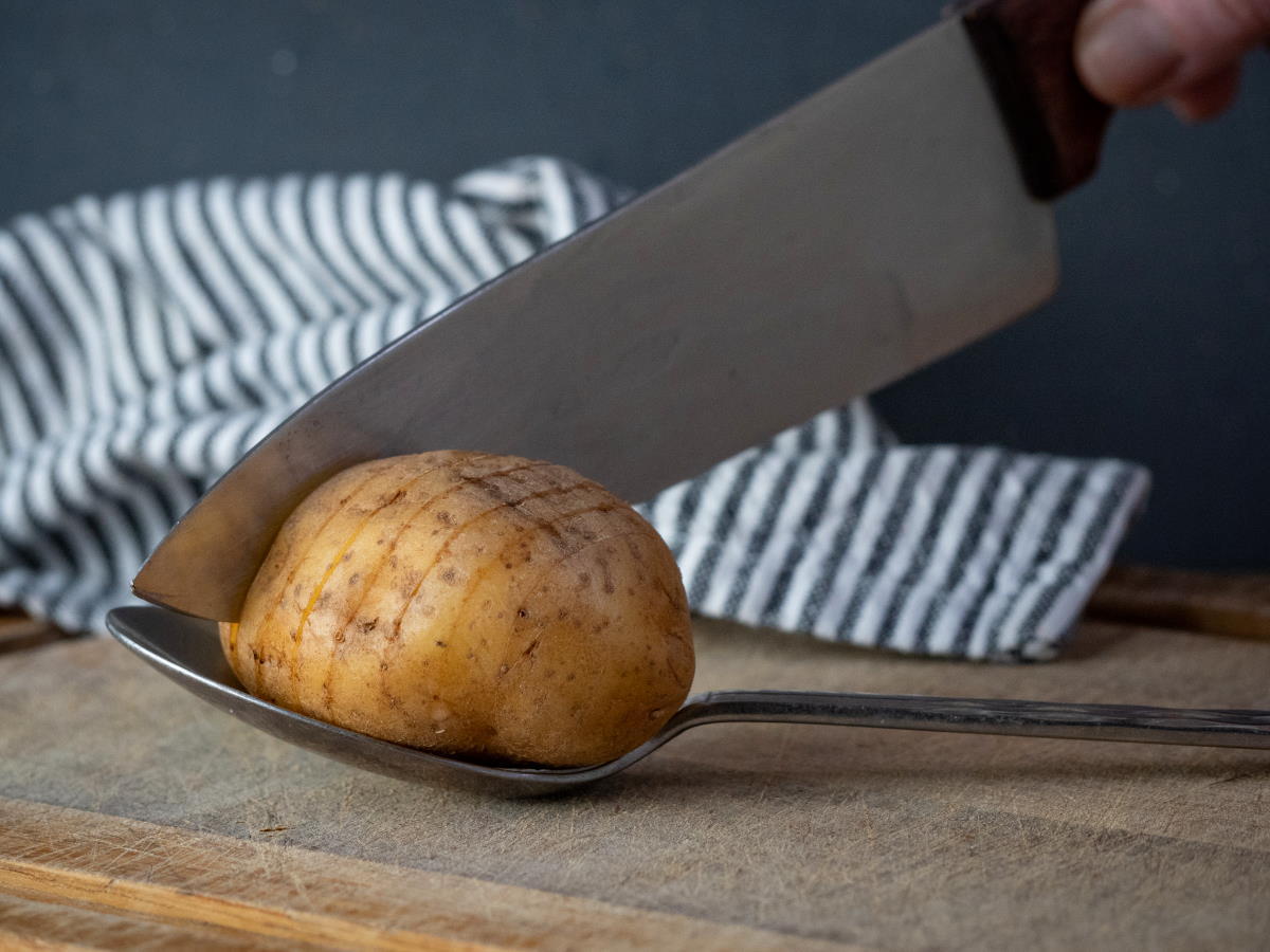 A potato on a spoon with a knife, on wooden cutting board.