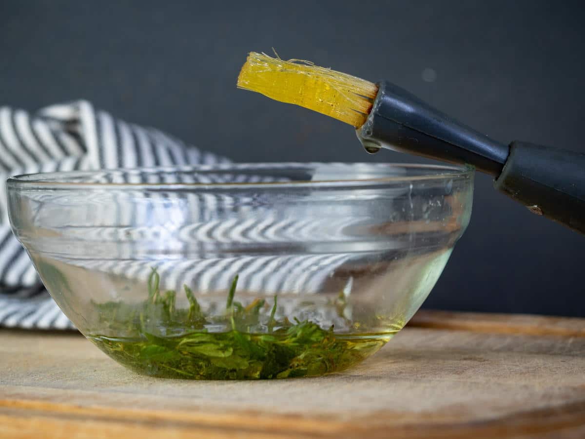 Bowl of olive oil and herbs with a silicone brush on wooden cutting board.
