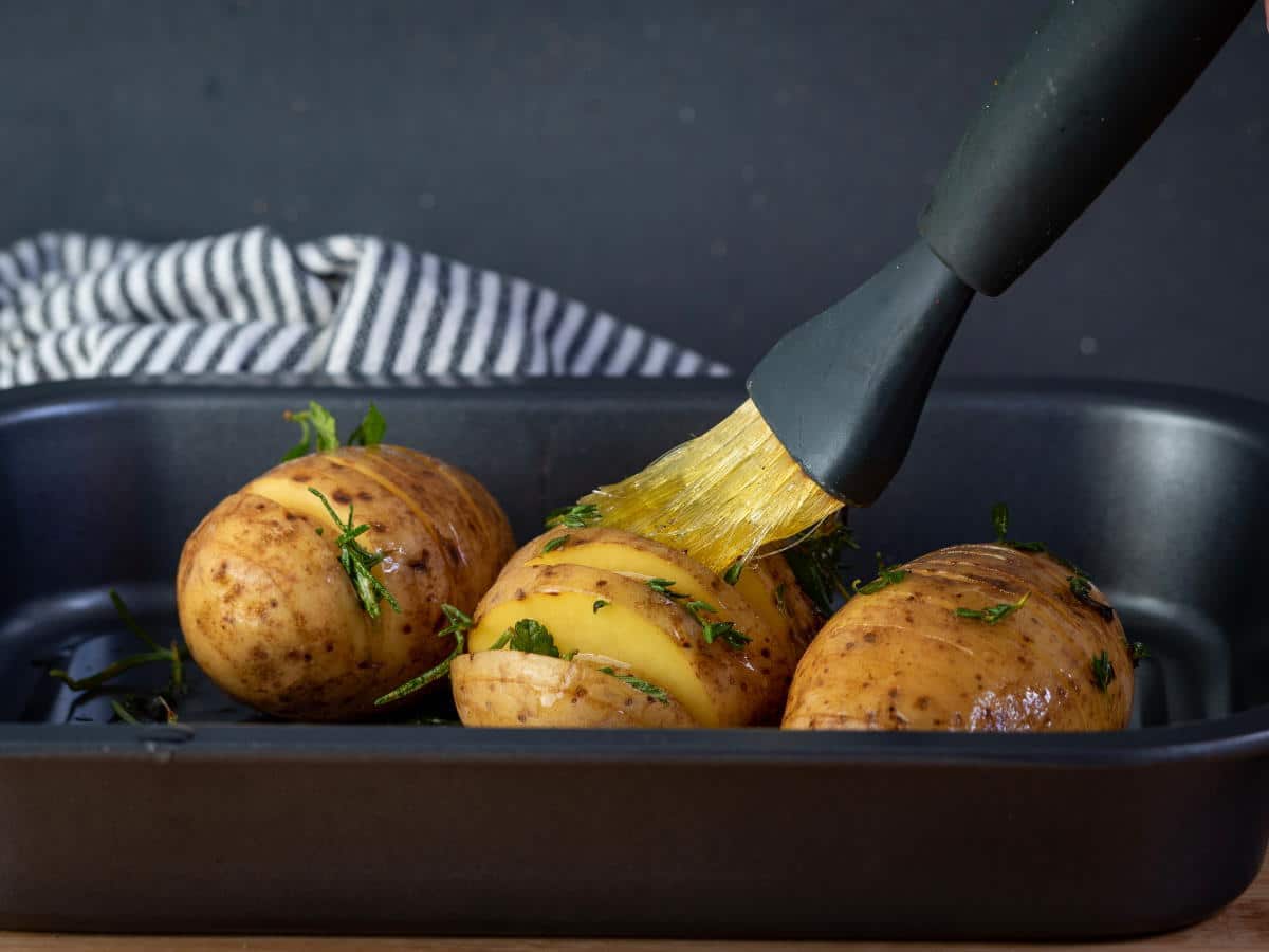 Hasselback potatoes with seasonings in baking dish and a silicone brush with olive oil.