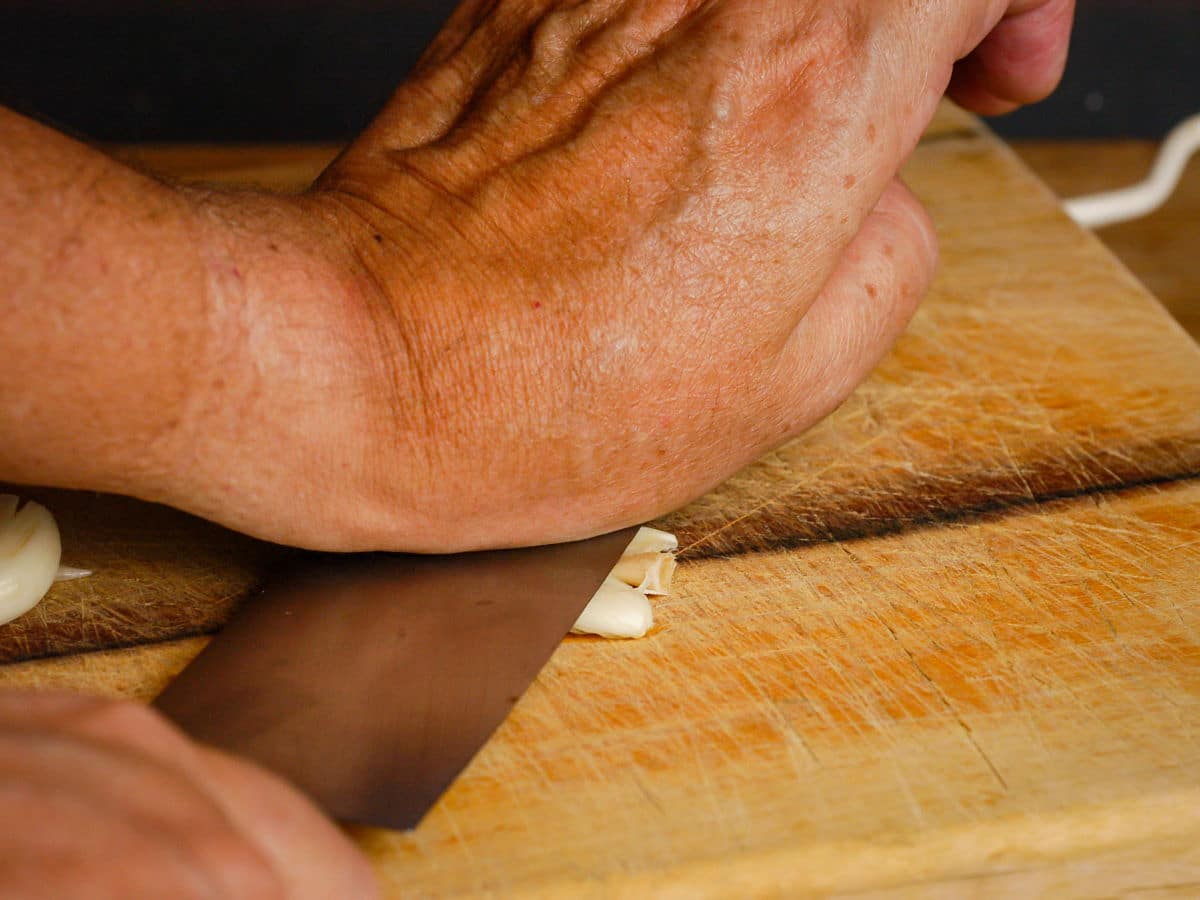 Garlic clove pressed under chef's knife with woman's hand.