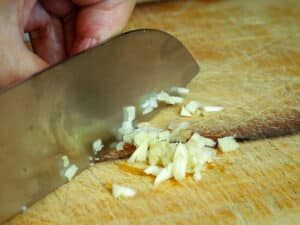 Chef's knife chopping garlic on wooden cutting board.