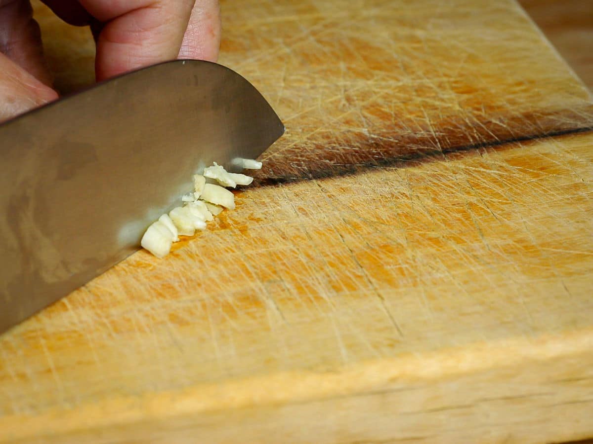 Chef's knife chopping garlic on wooden cutting board.