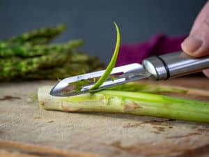 Asparagus stems getting peeled on wooden cutting board.