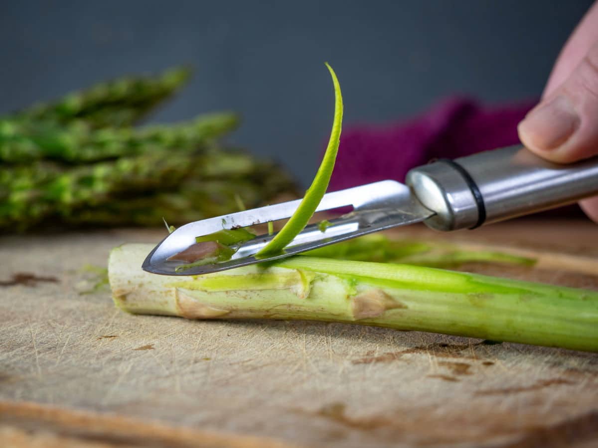 Asparagus stems getting peeled on wooden cutting board.