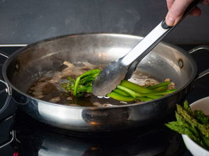 Asparagus in frying pan with metal tongs.