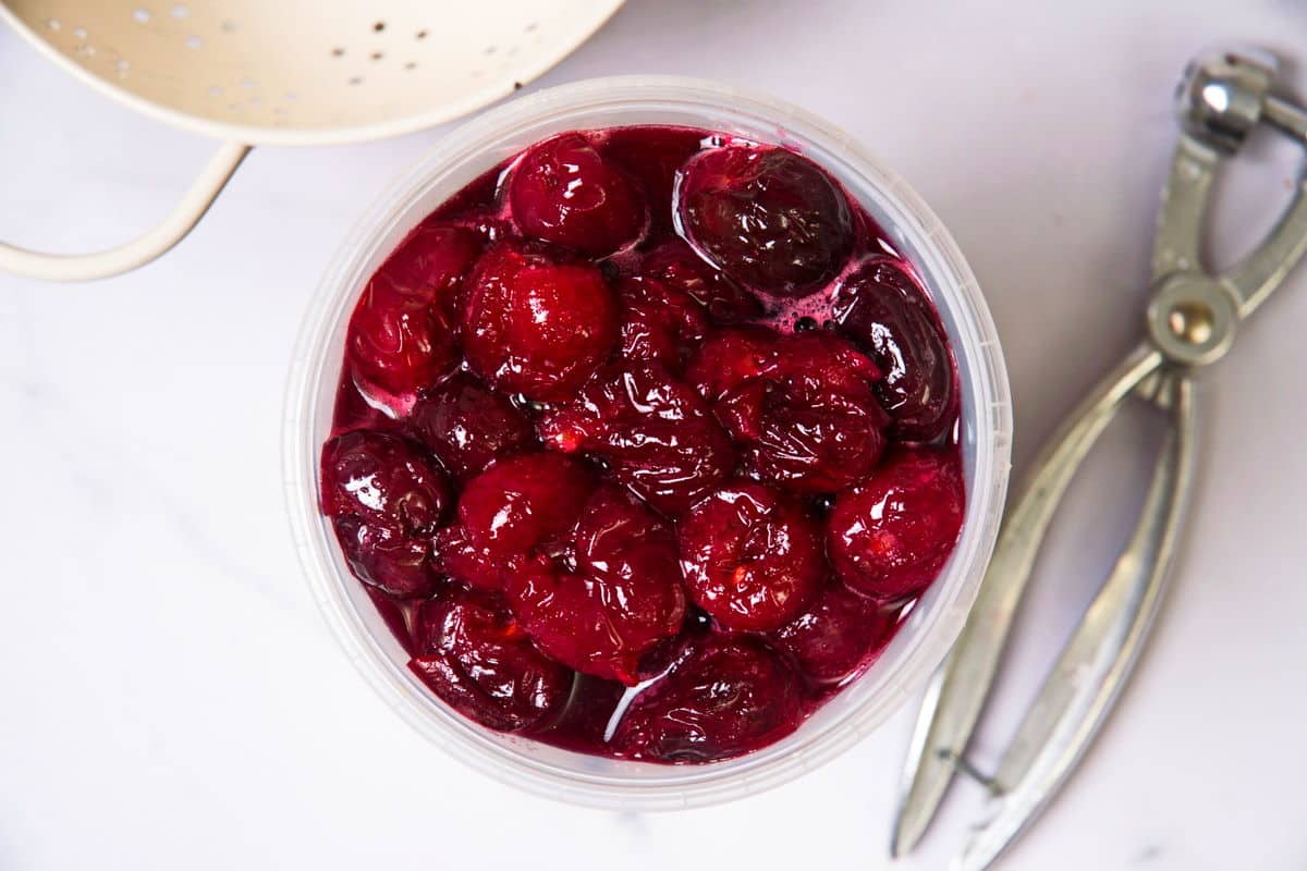 Cherries in a sugar syrup in plastic container, cherry pitter on the side.