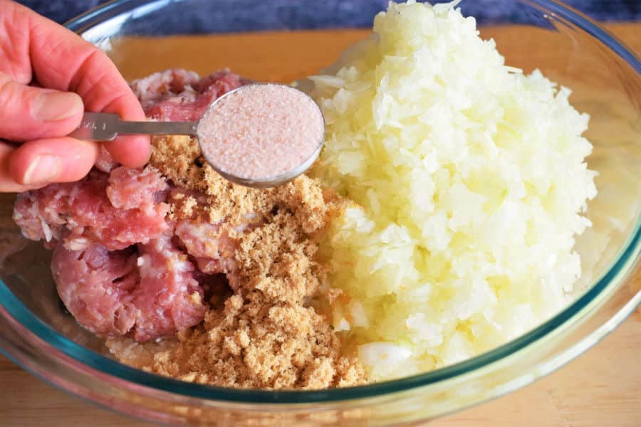 Clear glass bowl with cabbage roll ingredients on wooden table with a metal tablespoon of salt.