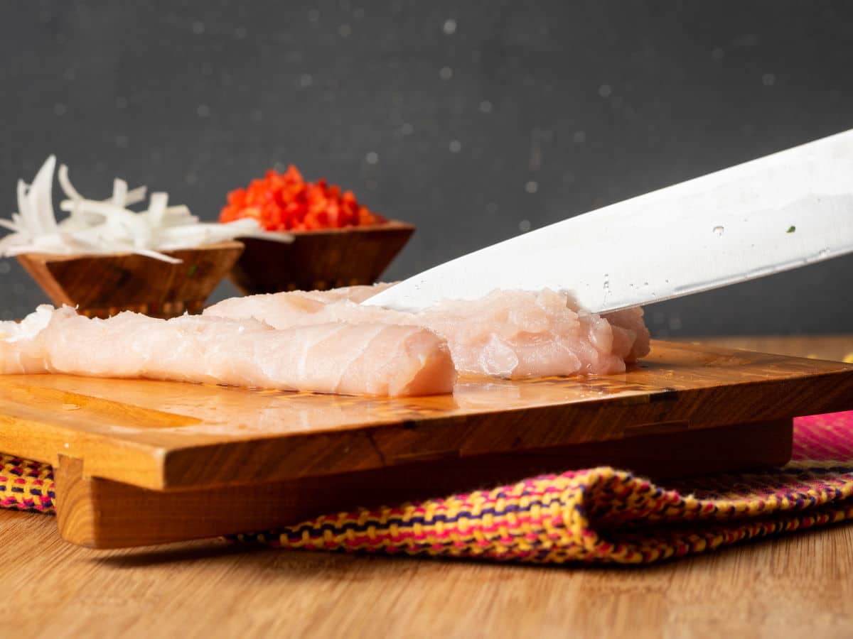 A knife slicing into raw white fish on cutting board.