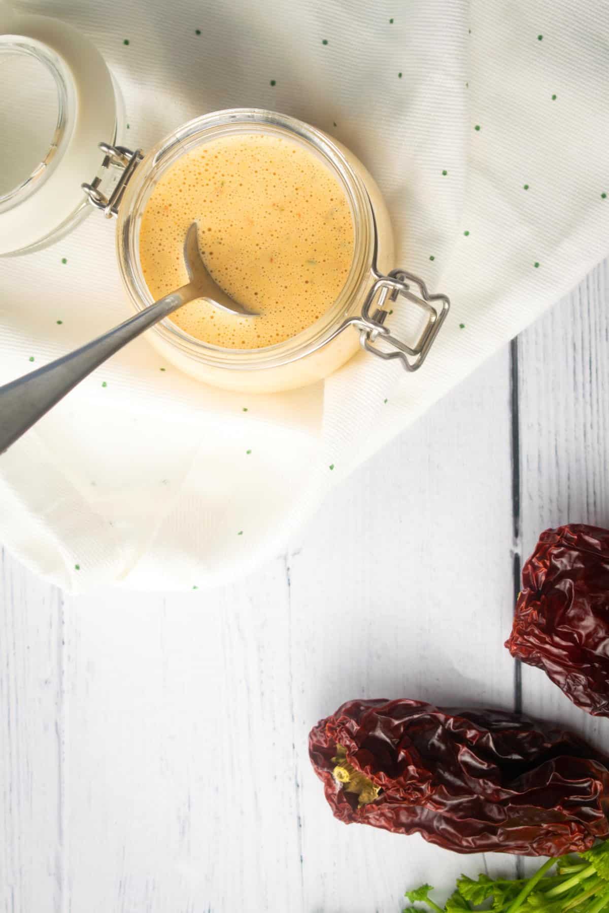 Chipotle sauce in a jar with a spoon, top view, on white wooden background. Chipotle peppers on the side.
