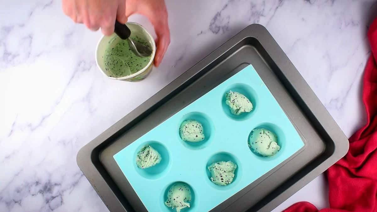 Mint ice cream balls in a muffin tray with a small tub of ice cream and a scoop on the side, marble background.