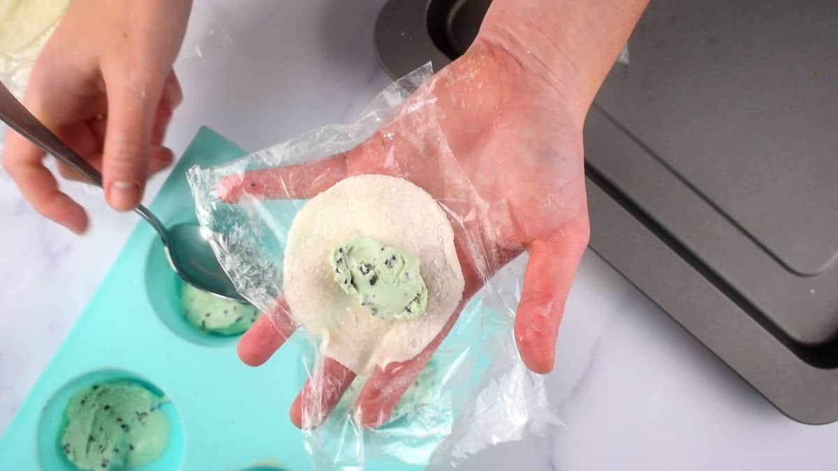 Mochi dough circle with mint chocolate chip ice cream on plastic wrap in a hand.