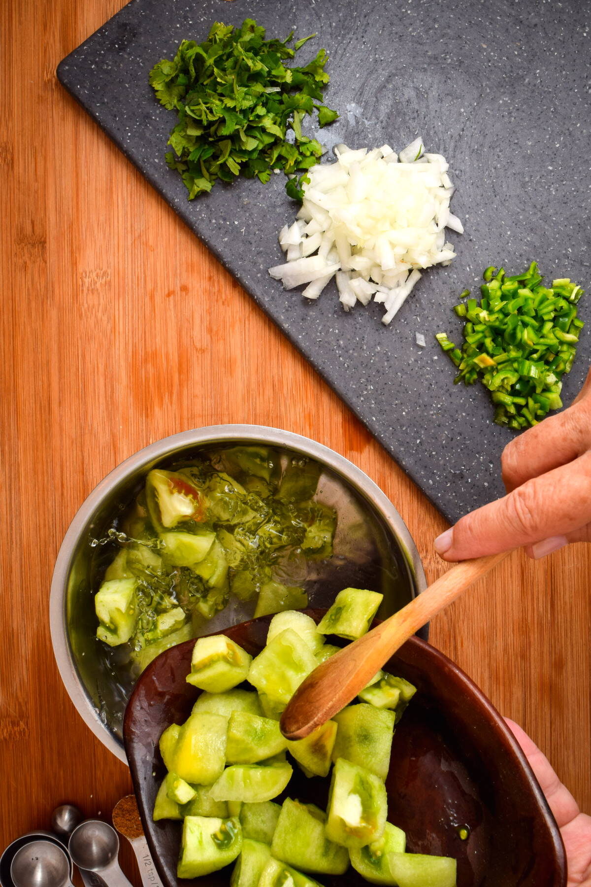 Pot of cooked tomatillo salsa ingredients and a wooden spoon. A cutting board with diced onion, green pepper and cilantro on the side.
