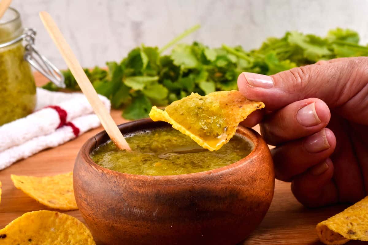 A wooden bowl of tomatillo salsa, a jar of tomatillo salsa, cilantro and tortilla chips in the background. A hand dipping a tortilla chip in the salsa.