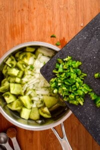 Diced green peppers on cutting board over pot of water, onions and tomatillos.