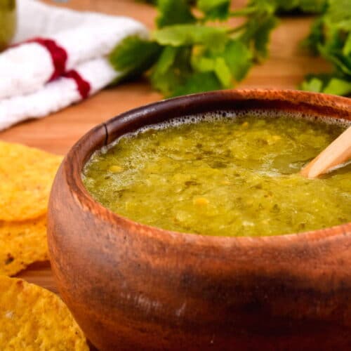 A wooden bowl of tomatillo salsa, a jar of tomatillo salsa, cilantro and tortilla chips in the background.