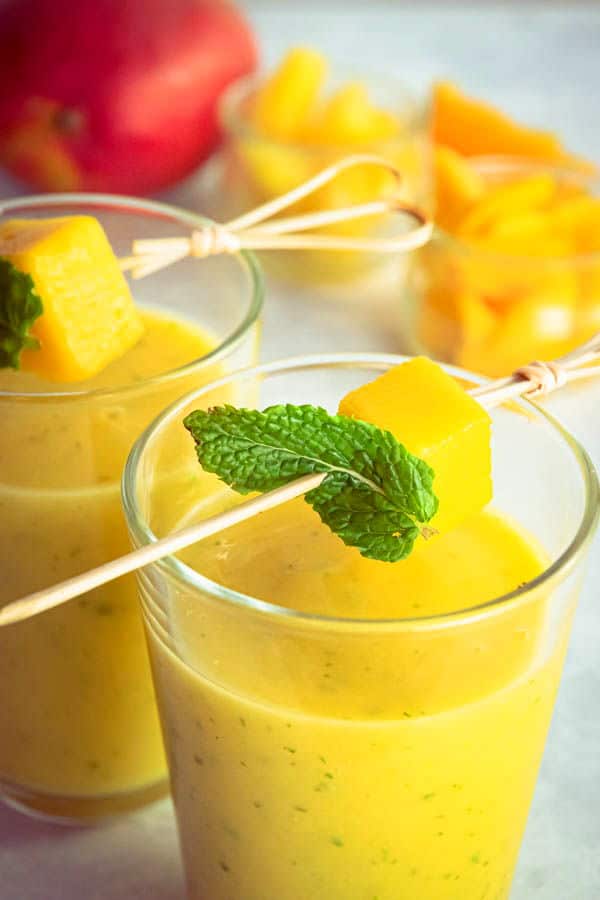 Mango pineapple smoothies in 2 glasses with mango and mint skewers, mango chunks in the background.