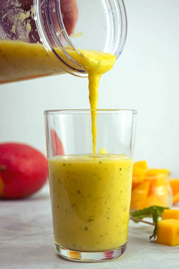 Mango pineapple smoothie being poured into a glass, fruit in the background.