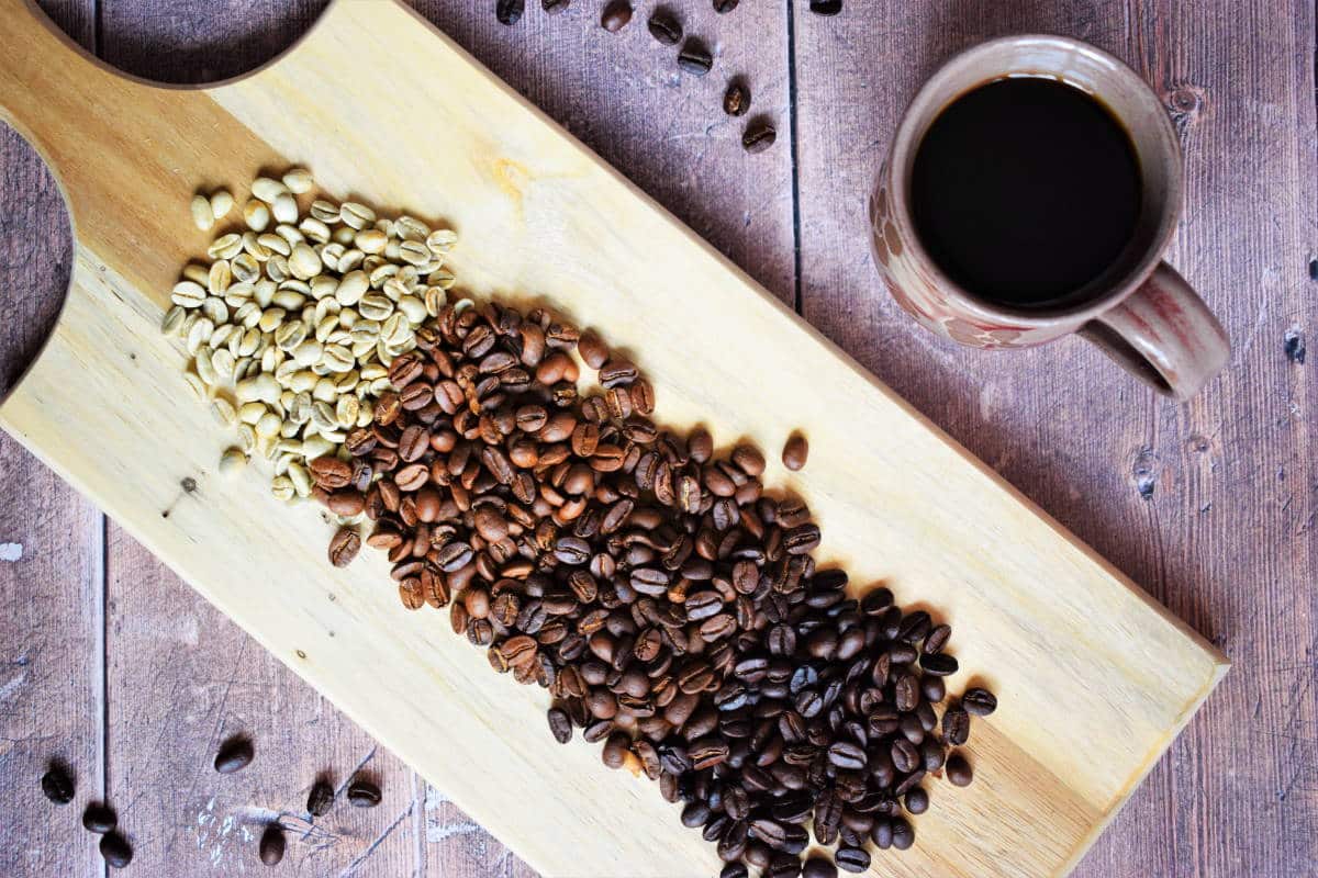 Green and brown coffee beans on wooden serving board, coffee mug on the side.