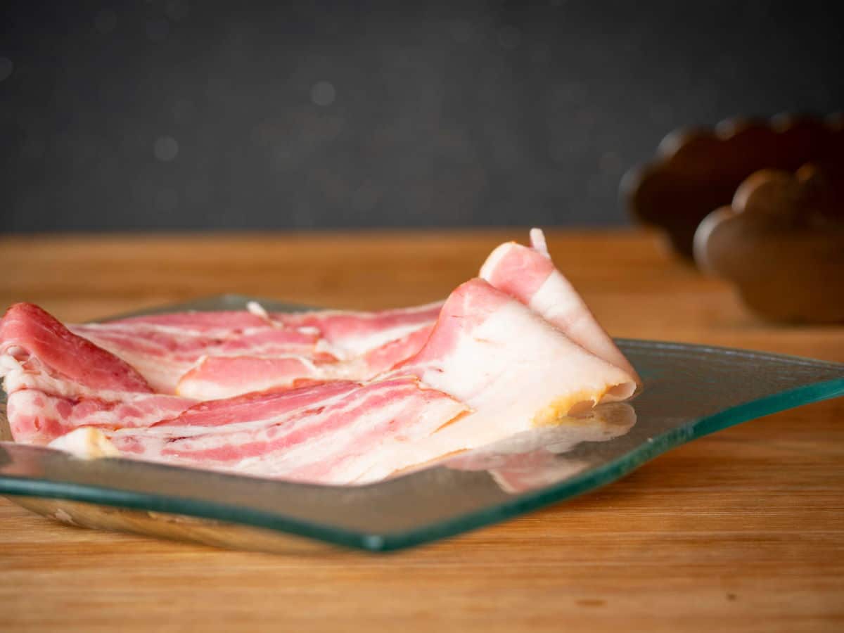 Raw bacon on a clear, glass plate, wooden background.