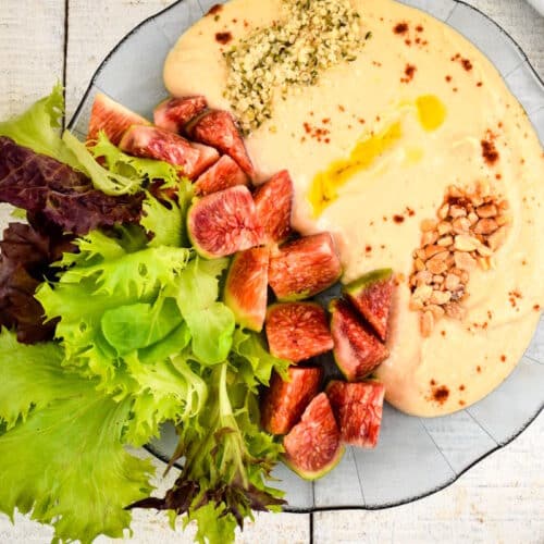 Top view of hummus dip in a bowl with fresh lettuce, figs, nuts, hemp seeds and olive oil.