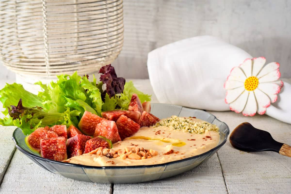 Hummus dip in a bowl with fresh lettuce, figs, nuts, hemp seeds and olive oil.