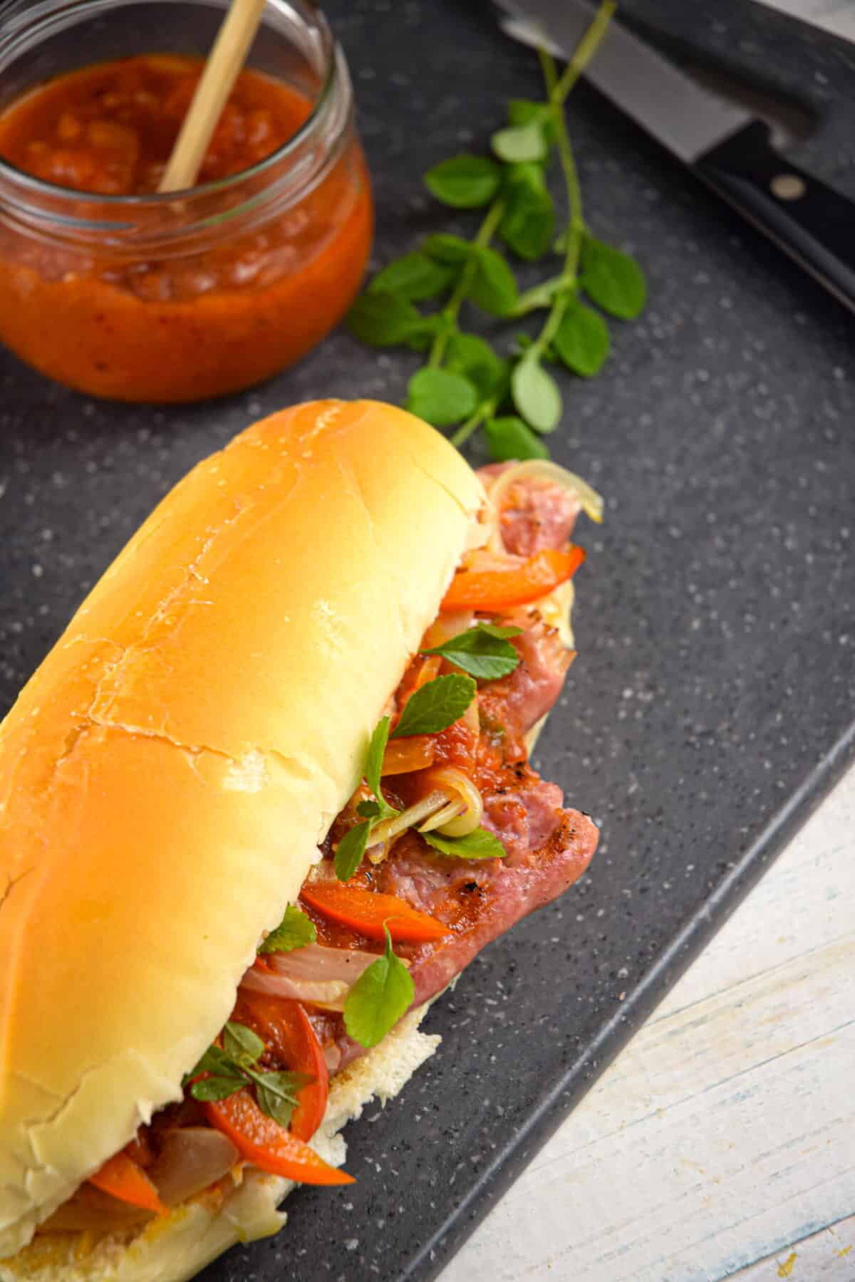 Italian sausage sandwich with fresh oregano and a jar of pepper sauce on dark cutting grey board.