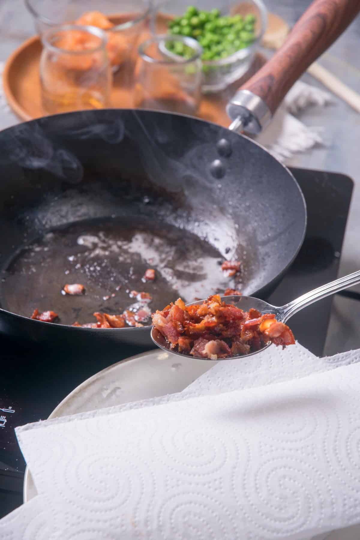 Cooked bacon bits in a spoon over paper towel, pan in the background.