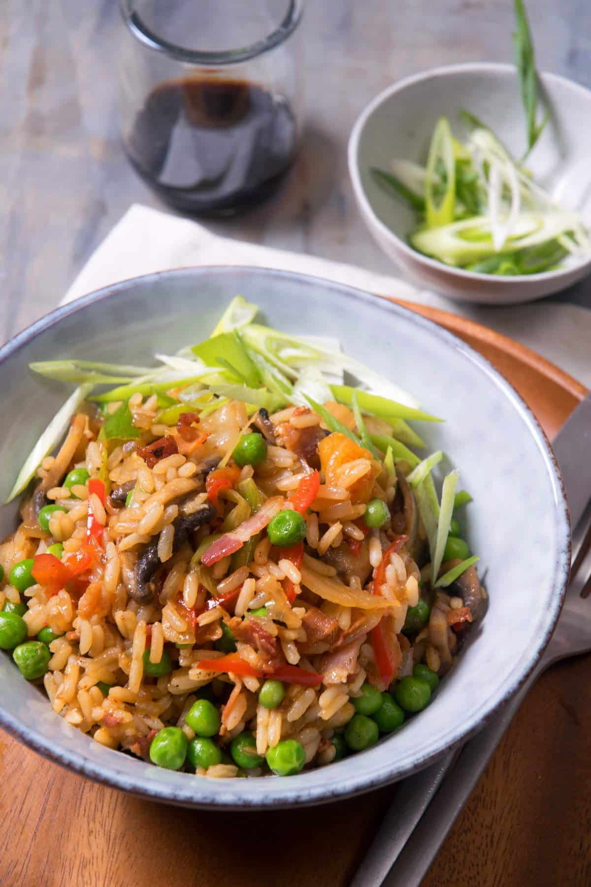 Kimchi bacon fried rice in bowl, with fork and knife on the side, sliced green onions in the background.