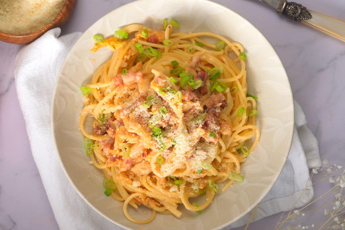 Kimchi carbonara in a white bowl with sliced green onions and grated Romano cheese.