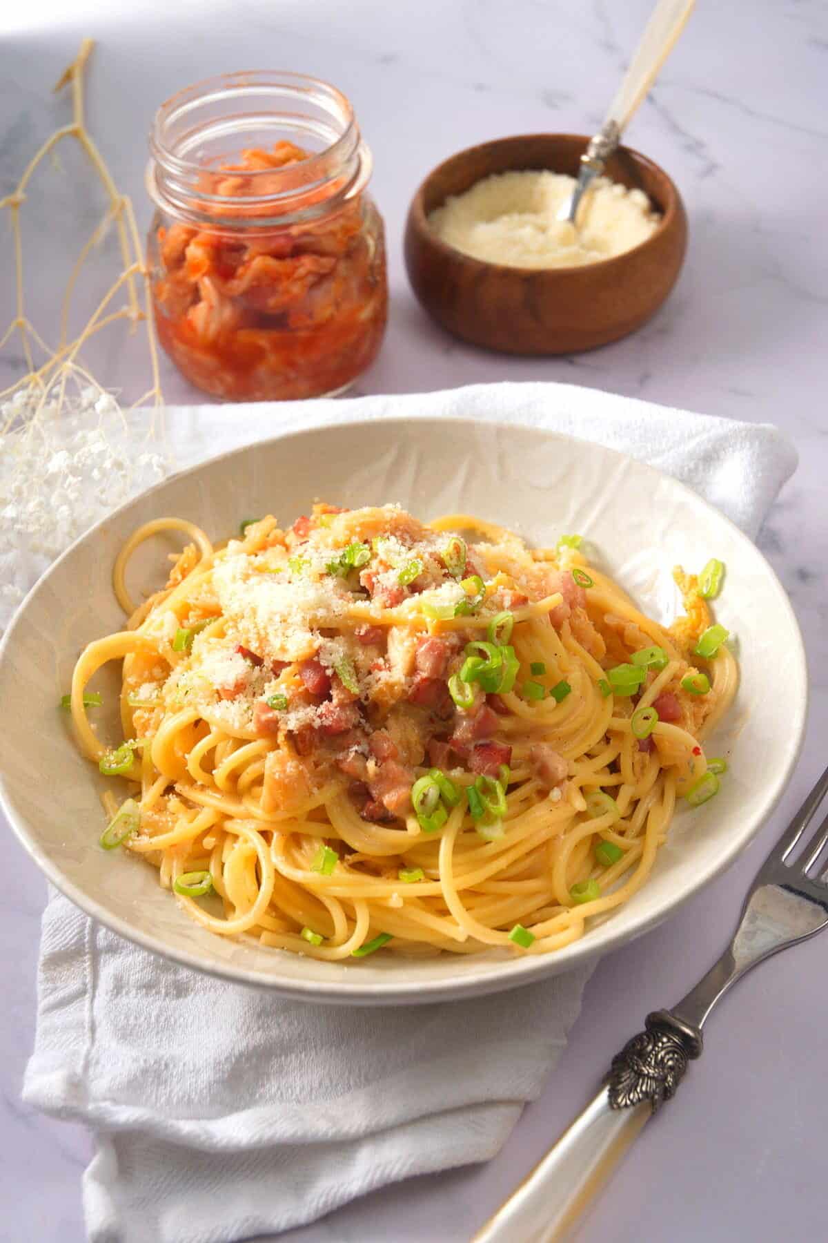 Kimchi carbonara in a white bowl with kimchi and grated pecorino in the background.