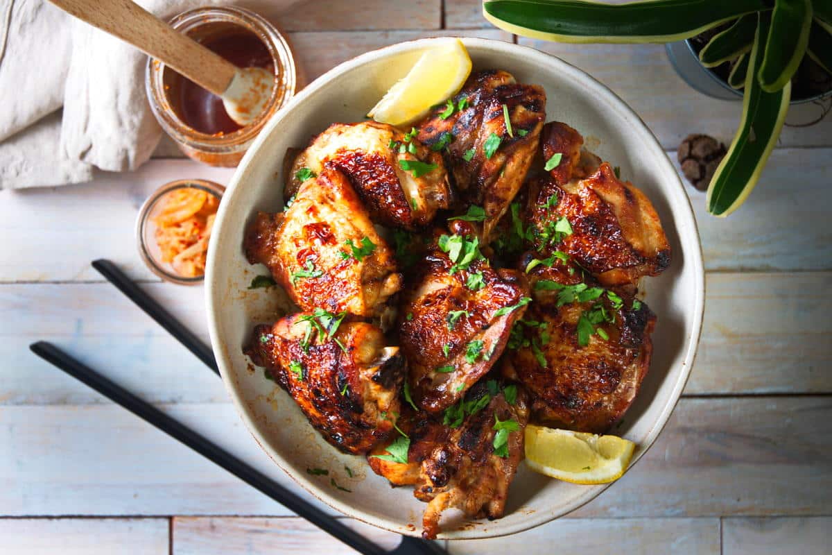 Top view of kimchi chicken thighs and lemon wedges in light dish. Chopsticks and kimchi on the side on wooden background.