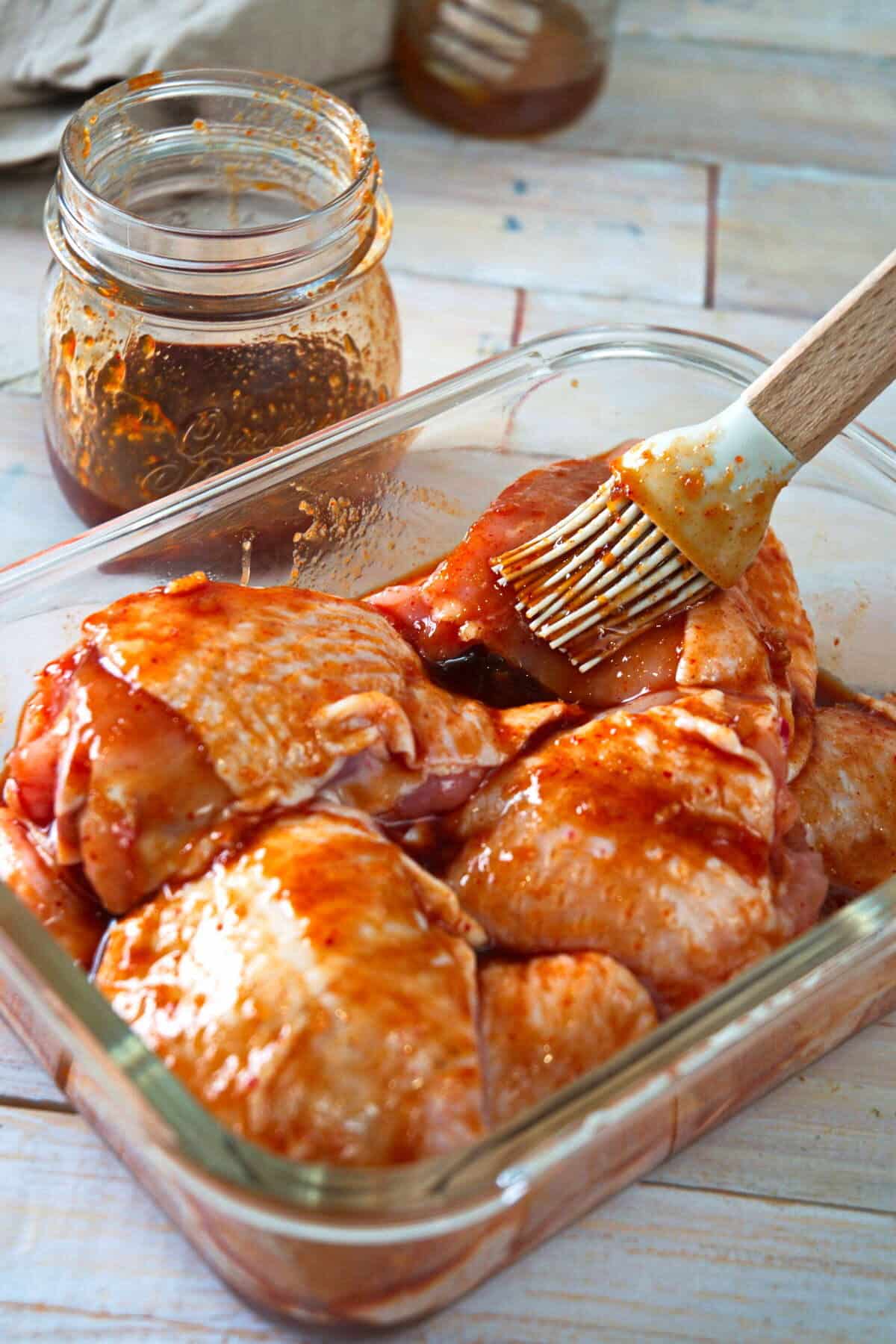 Chicken thighs in a marinade in glass container with silicone brush, wooden background.