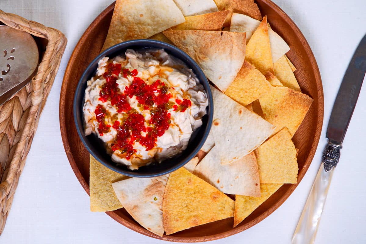 Top view of kimchi cream cheese spread in a small black bowl, surrounded by tortilla chips on a wooden plate.