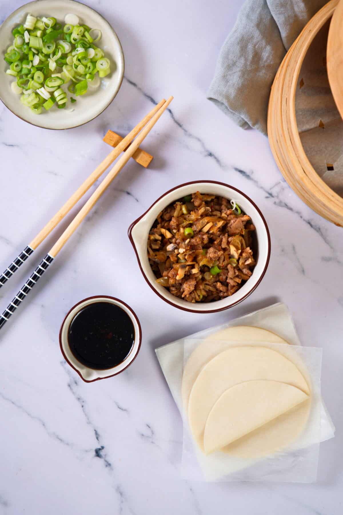 Kimchi dumpling ingredients prepped on marble background with a bamboo steamer in the corner.