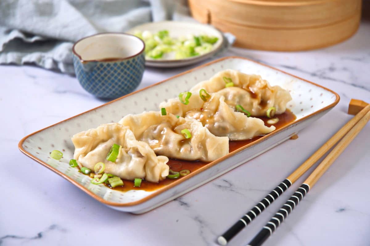 Kimchi dumplings with sauce and sliced green onions on a white and orange dish, chopsticks on the side.