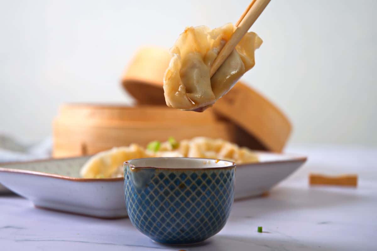 A dumpling held up in chopsticks over a small bowl of dipping sauce.