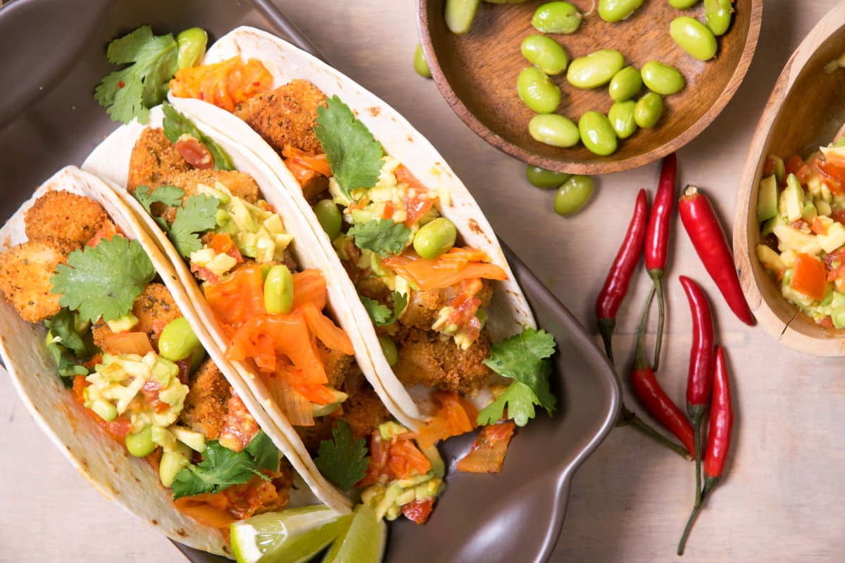 Kimchi and cod tacos in a brown dish, edamame beans and chili peppers on the side.