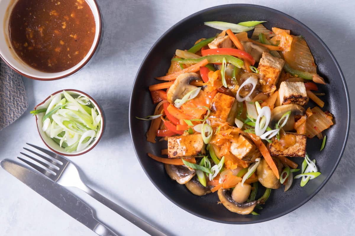 Kimchi tofu stir fry on black dish with a fork and knife, bowl of scallions and sauce on the side.