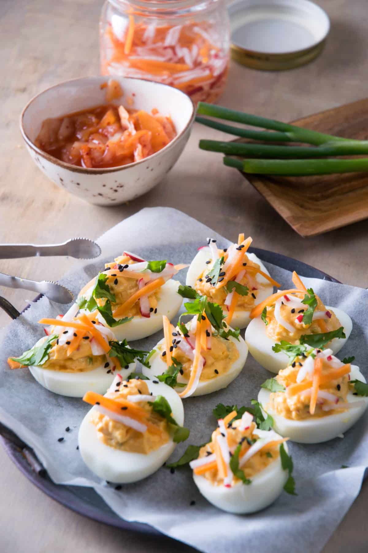 Kimchi deviled eggs on a plate with sliced green onion garnish, kimchi in a bowl in the background.
