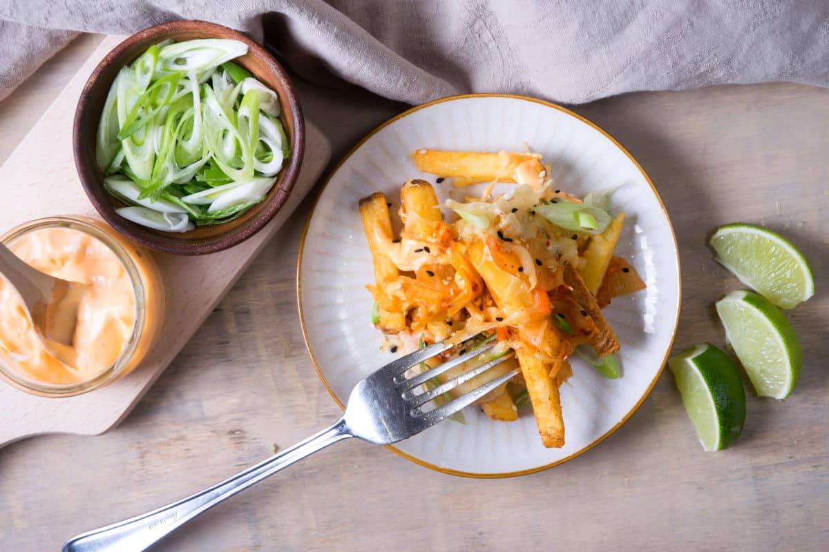 Fries with kimchi on a small plate. Lime wedges, sliced scallions and sriracha sauce on the side.