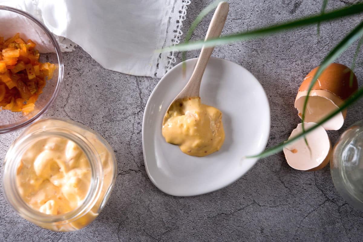 Kimchi mayo on a spoon on white dish, cracked egg shells and kimchi on the side, concrete look background.