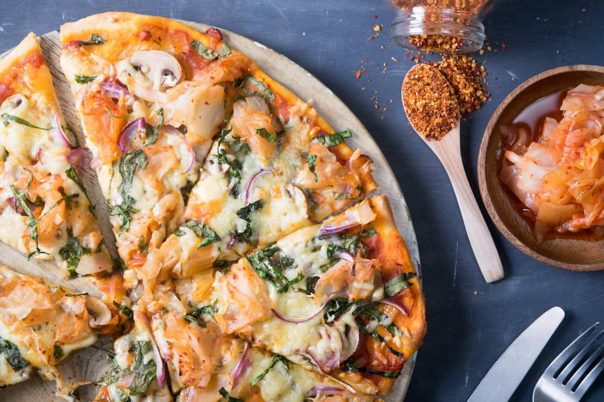 Sliced kimchi pizza on circular wooden cutting board, kimchi and chili pepper flakes on the side.