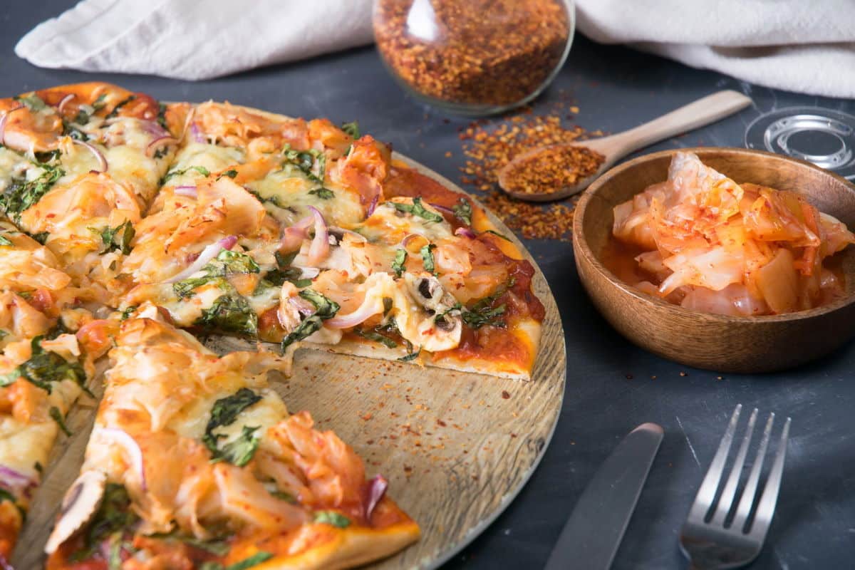 Sliced kimchi pizza on circular wooden cutting board, kimchi and chili pepper flakes on the side.